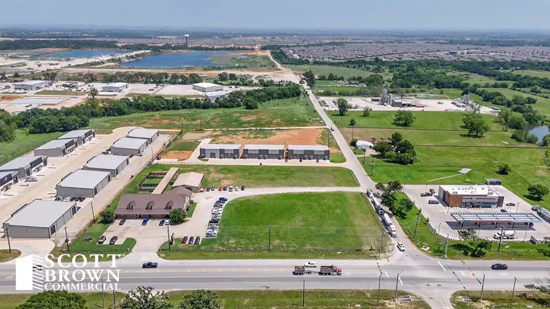 Aerial view over Stewart Road in Aubrey: light-industrial buildings and parking in the foreground, open land beyond, and a pond with a housing subdivision on the horizon.