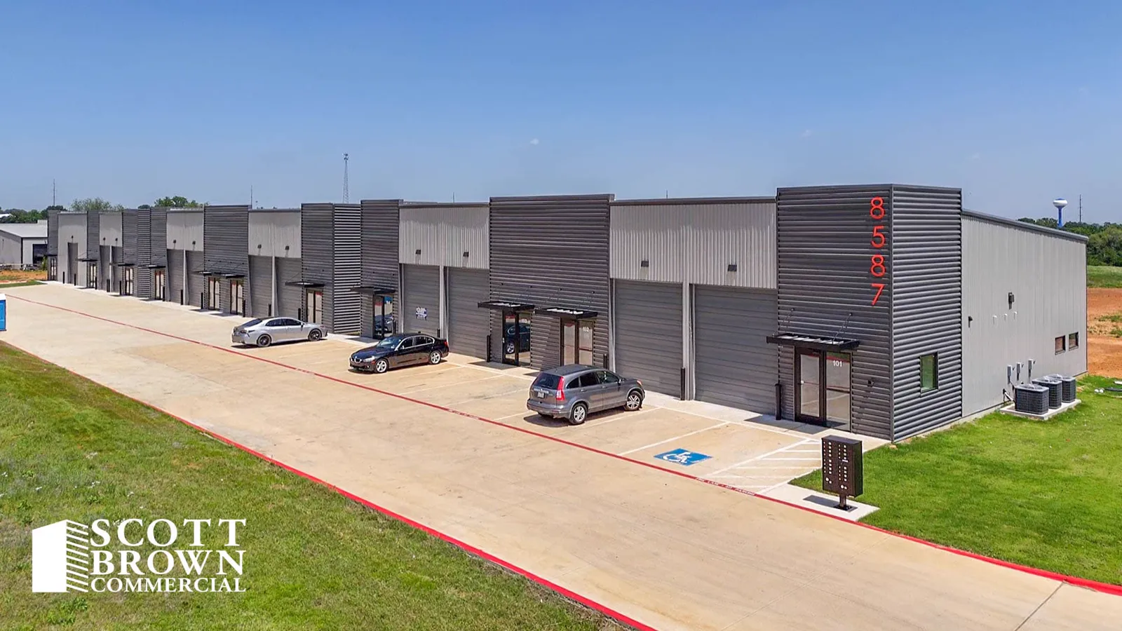 The front elevation of Fusion Business Park: a long terrace of flex suites in dark and light metal cladding, each with a grade-level overhead door, a glazed office entrance under an awning, and marked parking along a concrete apron.