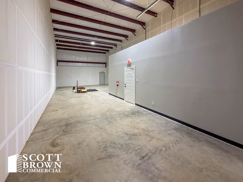 A warehouse bay at Fusion Business Park looking along the grey office partition, with red steel portal frames, white insulated metal walls, a sealed concrete floor, and a door through to the office.